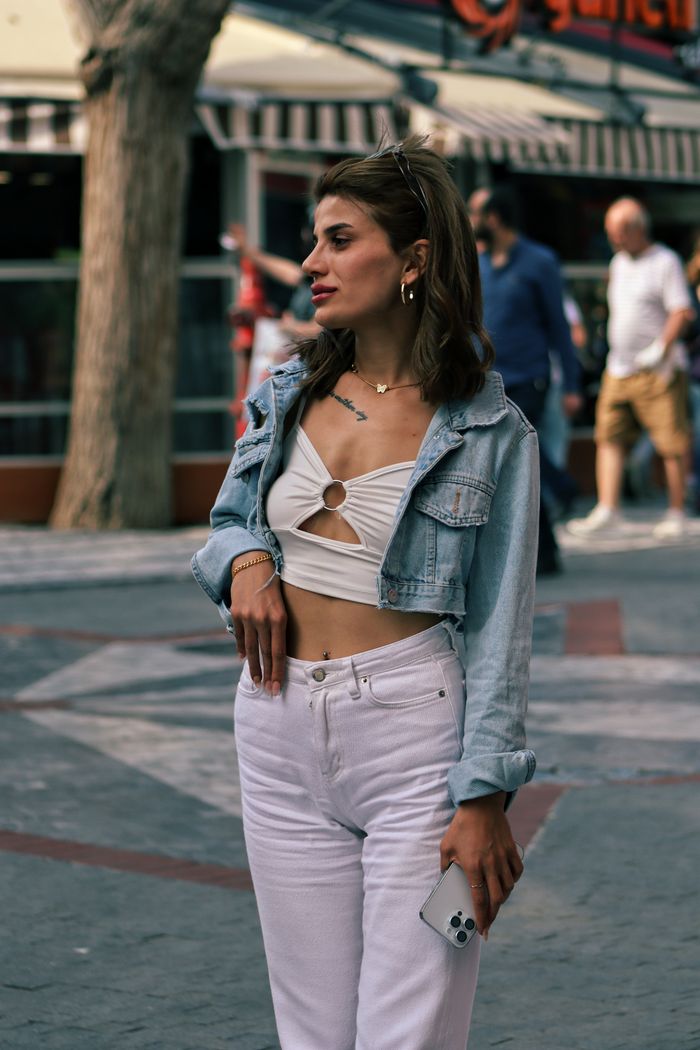 gallery-4 Stylish woman wears a jean jacket and holds a phone while standing outdoors on a city sidewalk.