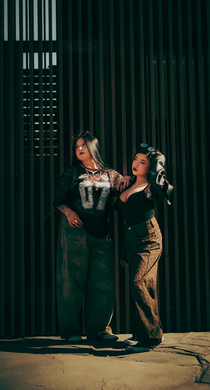 gallery-6 Stylish women pose in front of a slatted wall, showcasing urban fashion.